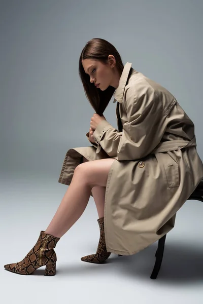 Full length of model in stylish trench coat sitting on chair and posing on grey - foto de stock