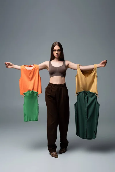 Full length of brunette model in crop top and trousers standing with outstretched hands and holding clothing on grey — Stockfoto