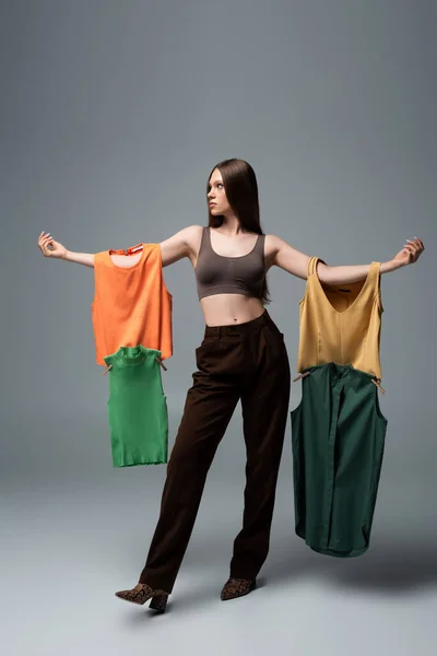 Full length of pretty model in crop top and trousers standing with outstretched hands while holding clothing on grey - foto de stock
