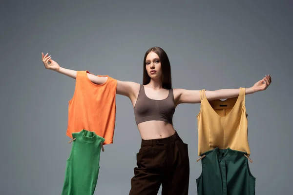 Pretty model in crop top and trousers standing with outstretched hands and holding clothing on grey — Stock Photo