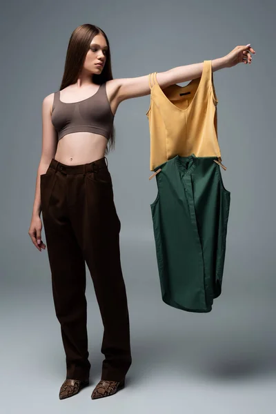 Full length of brunette model in crop top and trousers standing with outstretched hand and holding clothing on grey — Photo de stock