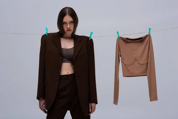 Brunette girl in brown suit pinned with clothespins on rope near long sleeve top isolated on grey — Stock Photo