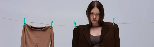 Brunette girl in brown suit pinned with clothespins on rope near long sleeve top isolated on grey, banner — Stockfoto