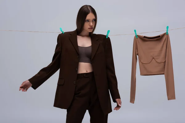 Teenage model in brown suit pinned with clothespins on rope near long sleeve top on grey — Fotografia de Stock