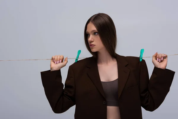 Stylish teenage model in brown blazer pinned with clothespins on rope posing isolated on grey — Stockfoto