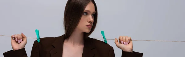 Stylish teenage model in brown blazer pinned with clothespins on rope posing isolated on grey, banner — Stock Photo