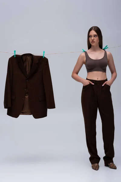 Stylish teenage girl in crop top pinned with clothespins on rope near brown blazer hanging on grey — Stockfoto