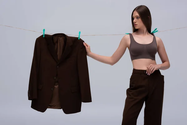 Stylish teenage model in crop top pinned with clothespins on rope touching brown blazer hanging isolated on grey — Stockfoto