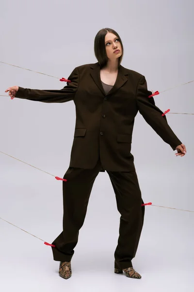 Full length of stylish teenage model in brown suit pinned with clothespins on rope posing on grey — Fotografia de Stock
