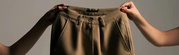 Обрезанный вид женщин, держащих переработанные бежевые брюки изолированные на сером, баннер — стоковое фото