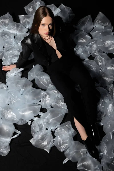 Высокий угол обзора брюнетки модели в стильном костюме позируя вокруг пластиковых отходов на черном — стоковое фото