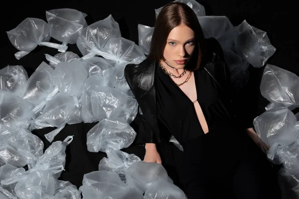 High angle view of pretty model in stylish suit posing around plastic waste on black — Fotografia de Stock