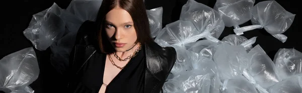 High angle view of brunette model in stylish suit posing around plastic waste on black, banner - foto de stock