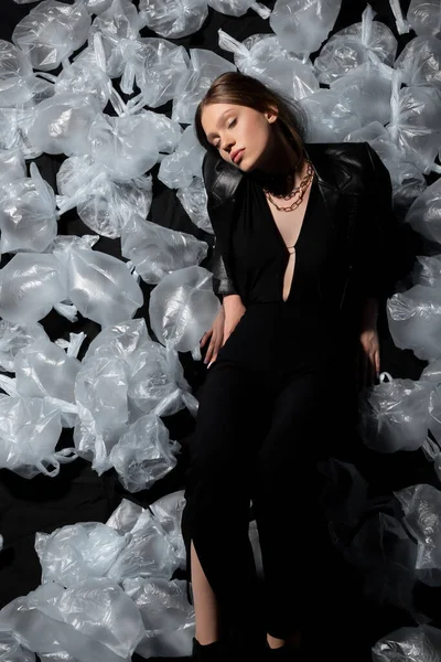Верхний вид брюнетки модели с закрытыми глазами в стильном костюме позирует вокруг пластиковых отходов на черном — стоковое фото