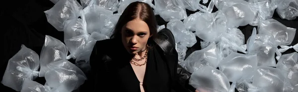 Top view of brunette model in stylish suit posing around plastic bags on black, banner — Stock Photo