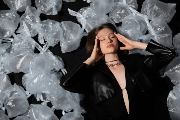 Top view of brunette model with closed eyes lying around plastic bags on black — Photo de stock