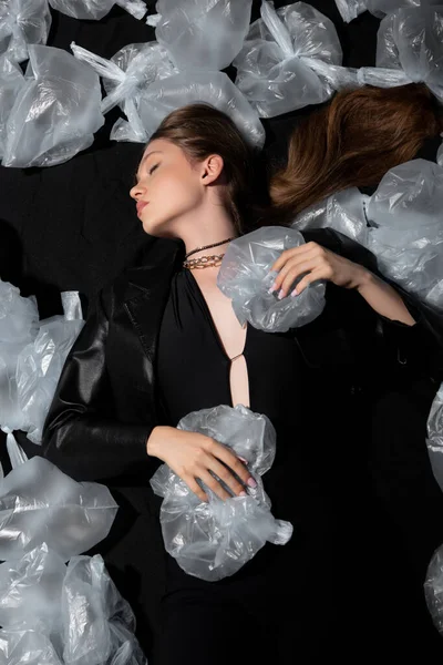 Top view of model with closed eyes lying around plastic bags on black — Photo de stock