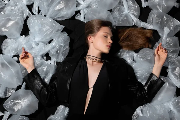 Top view of model with closed eyes lying around plastic garbage on black — Foto stock