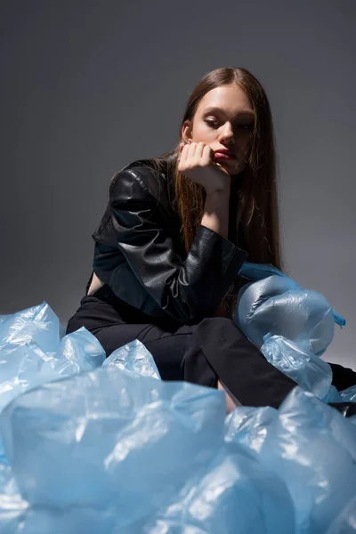 Sad model in stylish black suit posing around blue plastic bags on grey — Photo de stock