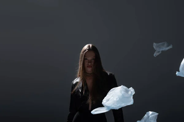 Брюнетка модель в стильному чорному костюмі позує біля літаючих пластикових пакетів на темно-сірому — стокове фото