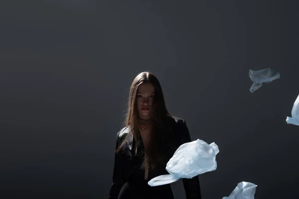 Brunette model in stylish black suit posing near flying plastic bags on dark grey — Stockfoto