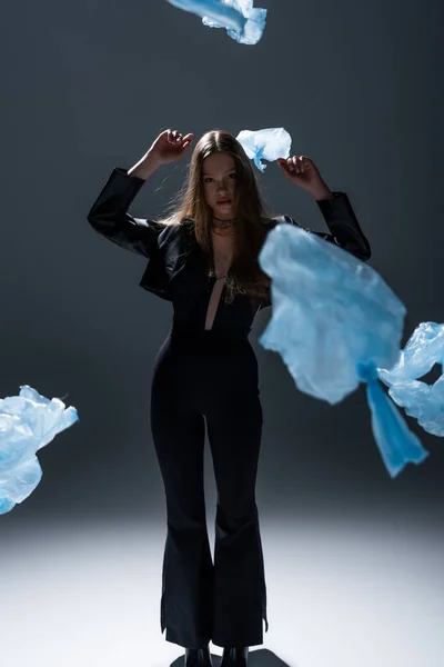 Full length of brunette model in stylish black suit posing near flying plastic bags on dark grey — Stock Photo