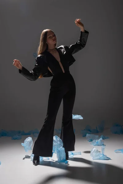 Full length of brunette model in stylish black suit gesturing near blue plastic bags on dark grey — Stock Photo