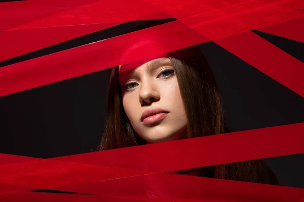 Модель-подросток, смотрящая на камеру через красные полоски из клейкой ленты, изолированной на черном, переработанной концепции моды — стоковое фото