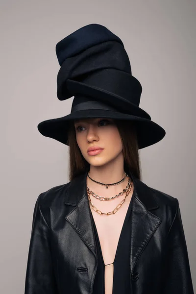 Brunette model in leather jacket and different black hats on head isolated on grey — Stockfoto