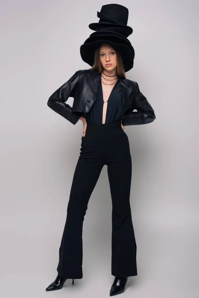 Full length of brunette model in leather jacket and different black hats on head posing on grey — Photo de stock