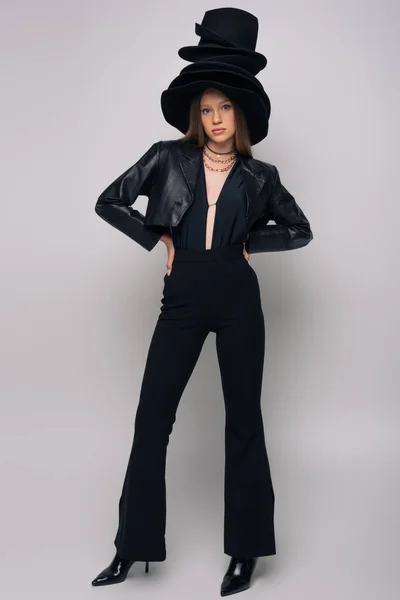 Full length of brunette model in leather jacket and different black hats on head posing on grey — Stock Photo