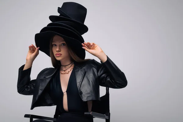 Teenage model in leather jacket and different black hats sitting isolated on grey — Stockfoto