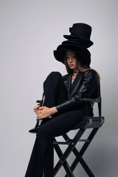 Brunette model in black leather jacket and different black hats sitting on chair on grey - foto de stock