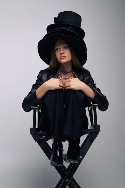 Model in black leather jacket and different black hats sitting on chair on gray - foto de stock