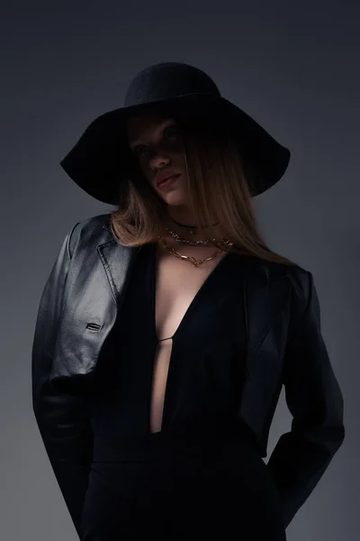 Стильна модель в капелюсі і чорній шкіряній куртці ізольована на сірому — стокове фото