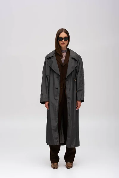 Повна довжина моделі в стильних сонцезахисних окулярах і модне траншеєподібне пальто позує на сірому — стокове фото