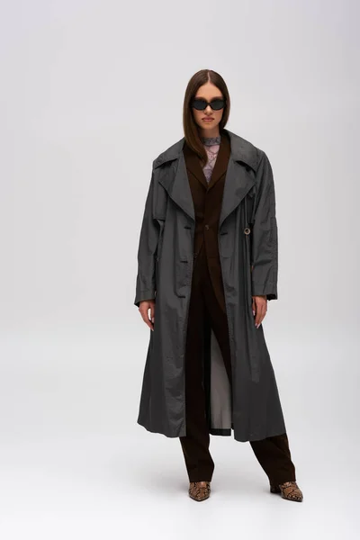 Повна довжина моделі в стильних сонцезахисних окулярах і траншеї пальто позує на сірому — стокове фото