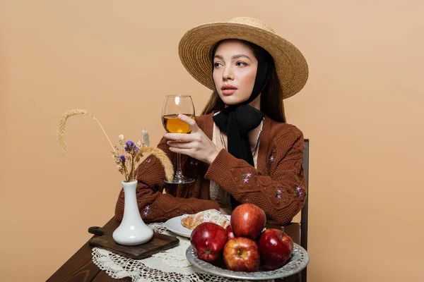 Stylish woman in sun hat holding wine near food on table isolated on beige — Foto stock