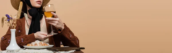 Cropped view of woman in sun hat holding glass of wine near plants and croissant on table isolated on beige, banner - foto de stock