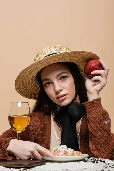 Молодая модель в соломенной шляпе держит яблоко и смотрит в камеру рядом с круассаном и вино изолированы на бежевый — стоковое фото