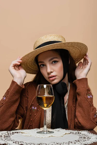 Trendy woman in straw hat looking at camera near glass of wine on table isolated on beige — Stockfoto