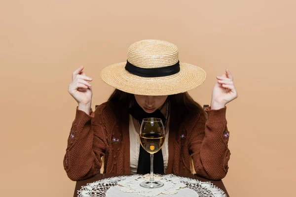 Молода жінка в сонячному капелюсі сидить біля келиха вина на столі ізольовано на бежевому — стокове фото