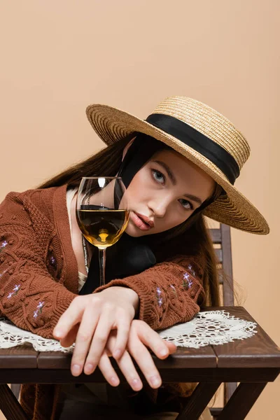 Брюнетка в солнечной шляпе смотрит в камеру рядом с бокалом вина, изолированного на бежевом — стоковое фото