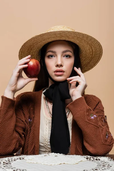 Портрет молодой женщины в соломенной шляпе, держащей яблоко возле стола, изолированного на бежевом — стоковое фото