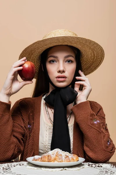 Portrait of woman in cardigan and sun hat holding apple near croissant isolated on beige — Stock Photo