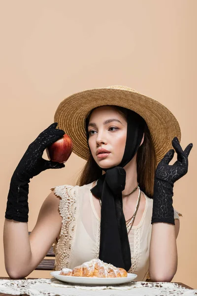 Модная женщина в соломенной шляпе и перчатках держит яблоко возле круассана изолированы на бежевый — стоковое фото