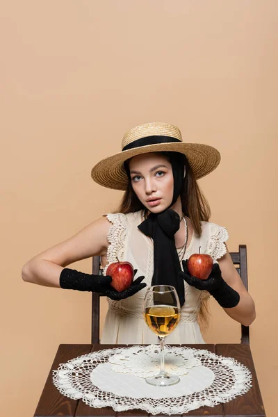 Fashionable woman in gloves and sun hat holding apples near wine isolated on beige — Foto stock