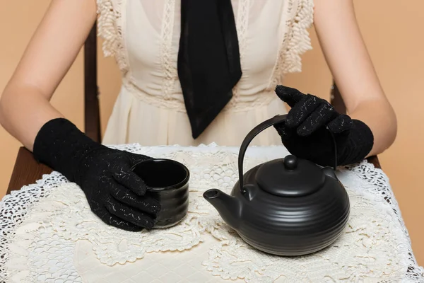 Обрезанный вид женщины в перчатках держа чашку и чайник на столе изолированы на бежевый — стоковое фото