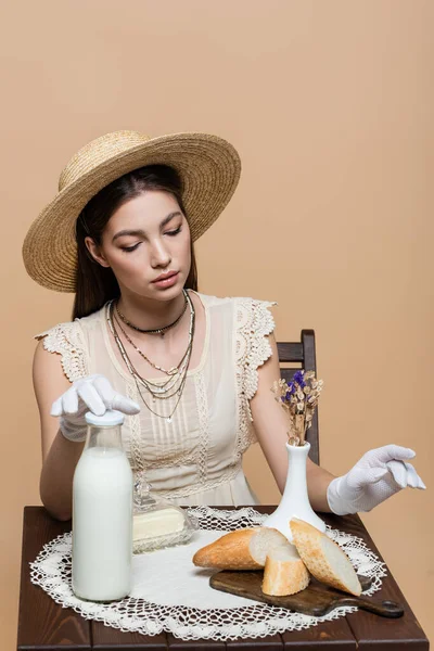 Pretty woman in sun hat and gloves sitting near bread and milk on table isolated on beige — Fotografia de Stock