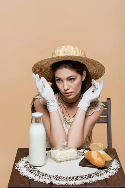 Молода жінка в рукавичках і сонячному капелюсі сидить біля пляшки молока і хліба ізольовані на бежевому — стокове фото
